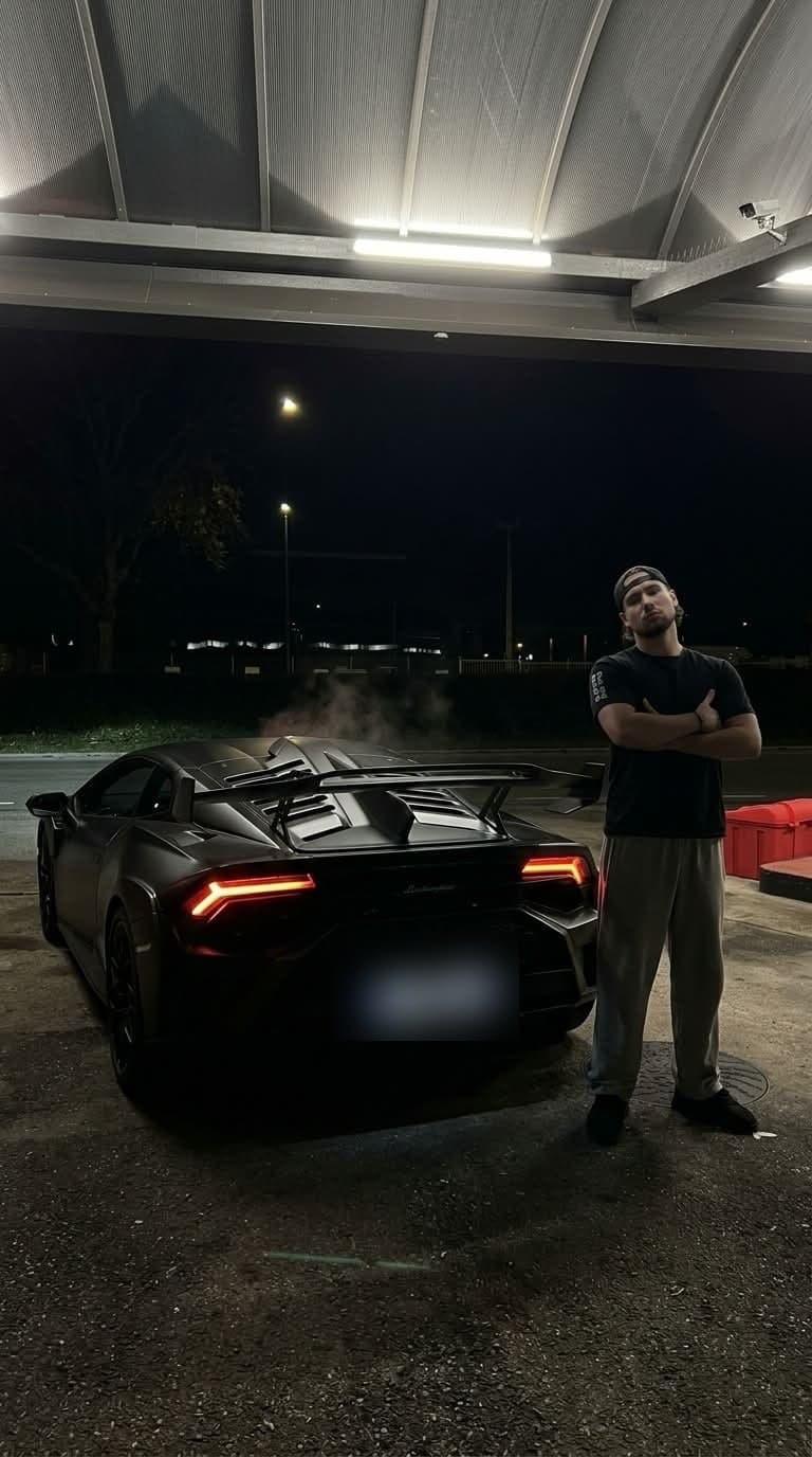 Luka at night, arms crossed, in front of his black Lamborghini under a gas station canopy